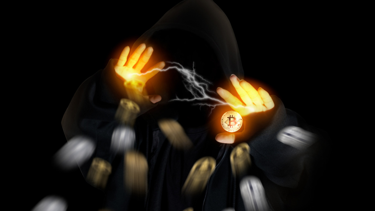 Hooded man in shadow with crypto currency bitcoin flying on black background.
