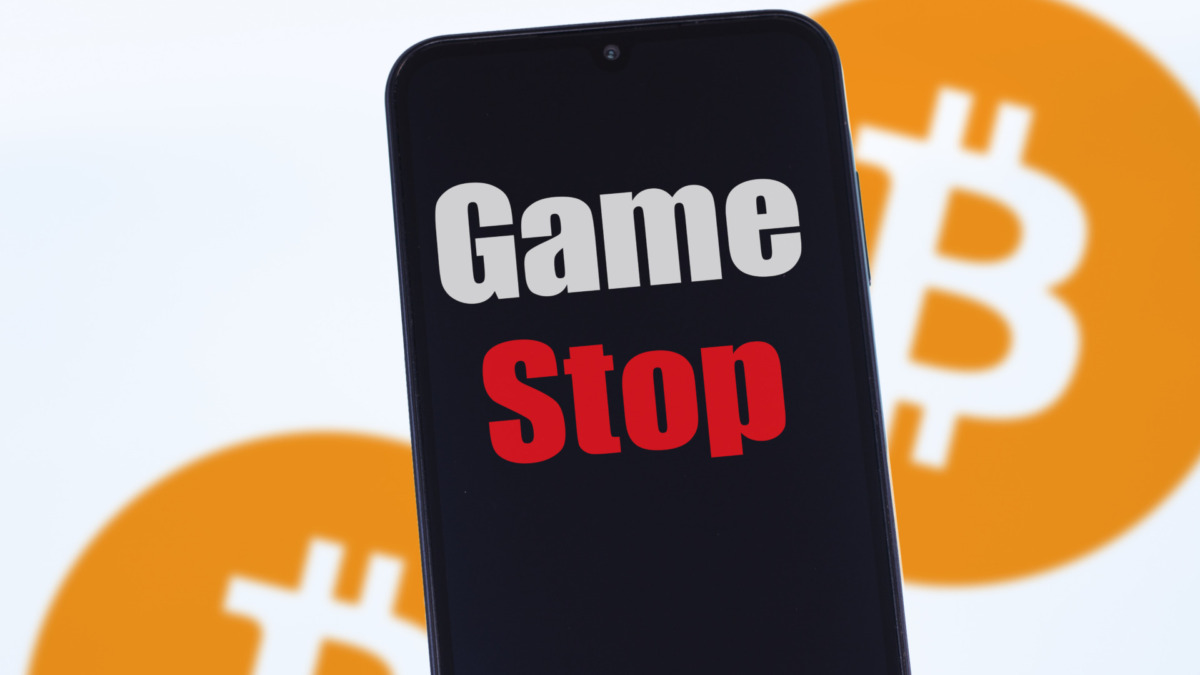 Close up on the logo gameStop, in the blurred background, two instances of the bitcoin logo