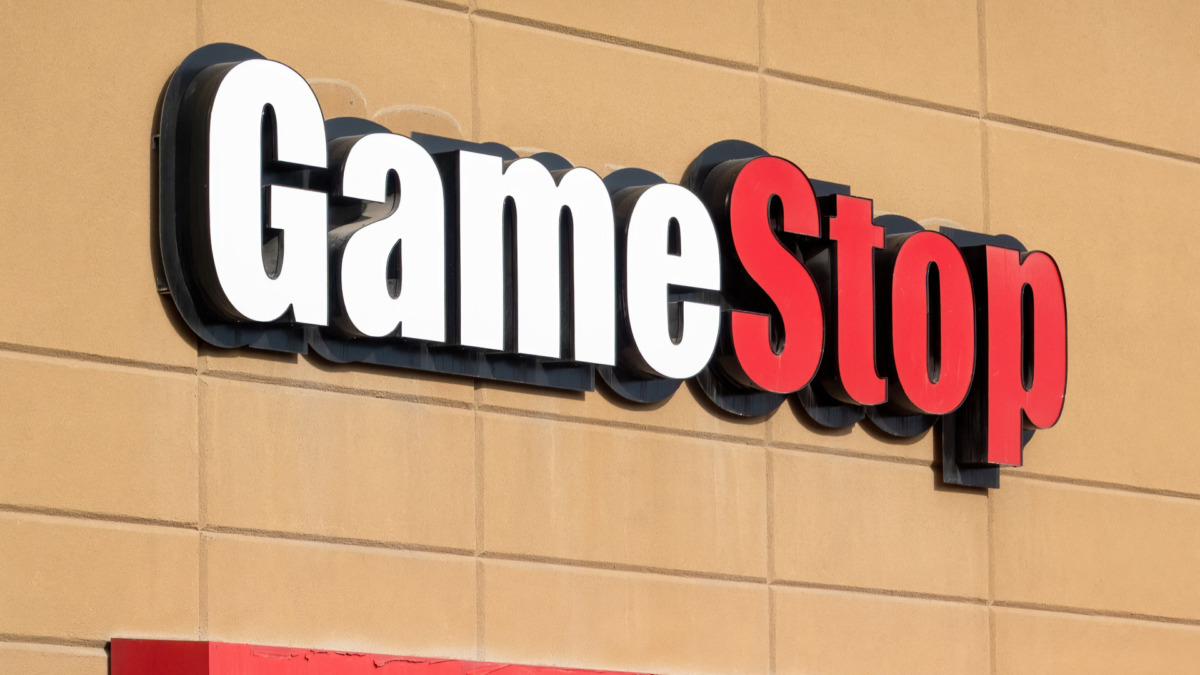 A close-up shows a GameStop store sign with white "Game" and red "Stop" lettering against a beige tiled building.