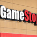 A close-up shows a GameStop store sign with white "Game" and red "Stop" lettering against a beige tiled building.