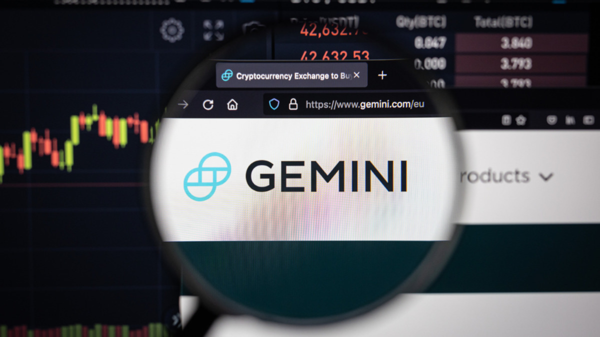 Gemini crypro exchange logo on a website with blurry stock market developments in the background, seen on a computer screen through a magnifying glass
