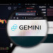 Gemini crypro exchange logo on a website with blurry stock market developments in the background, seen on a computer screen through a magnifying glass