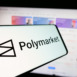 Smartphone with logo of crypto prediction market company Polymarket on screen in front of website. Focus on center-left of phone display.