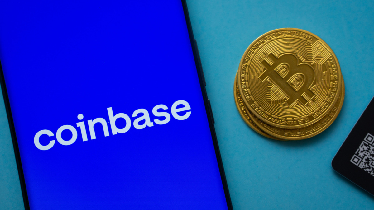 Coinbase logo on smartphone screen near Bitcoin coin and QR code on blue background.