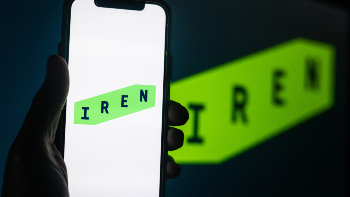 Silhouette of Hand Holding iPhone with IREN Logo in Front of Computer Screen Displaying Logo – Energy and Utilities Provider (IREN)