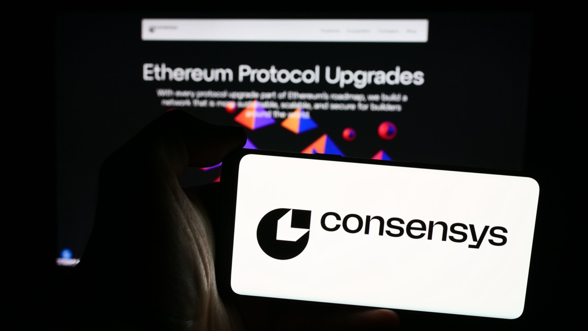 Person holding mobile phone with logo of American blockchain software company Consensys Inc. in front of web page. Focus on phone display.
