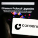 Person holding mobile phone with logo of American blockchain software company Consensys Inc. in front of web page. Focus on phone display.