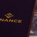 On screen mobile have logo of the binance trading website. And there are bnb coins and dollars laying nearby.