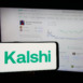 Person holding smartphone with logo of US prediction market company Kalshi Inc. on screen in front of website. Focus on phone display.