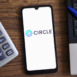 The Circle company logo on a smartphone screen against a background of a laptop, calculator, and pen.