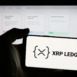 Person holding mobile phone with logo of cryptocurrency platform XRP Ledger (XRPL) on screen in front of web page. Focus on phone display.