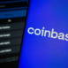 Coinbase logo is displayed on a smartphone screen, with the coinbase login page visible on a computer screen in the background.