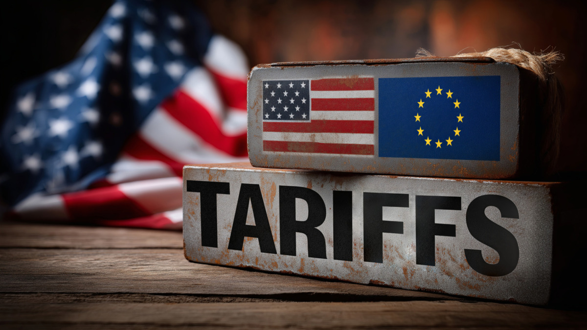 Two metallic blocks, one with the US flag and the other with the EU flag, are stacked on a wooden surface with a "TARIFFS" sign in front.