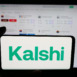 Person holding cellphone with logo of US prediction market company Kalshi Inc. on screen in front of webpage. Focus on phone display.